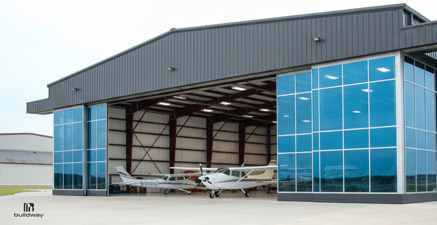 Large steel aircraft hangar with wide open doors, blue-tinted glass panels, and small airplanes parked inside on a concrete apron.