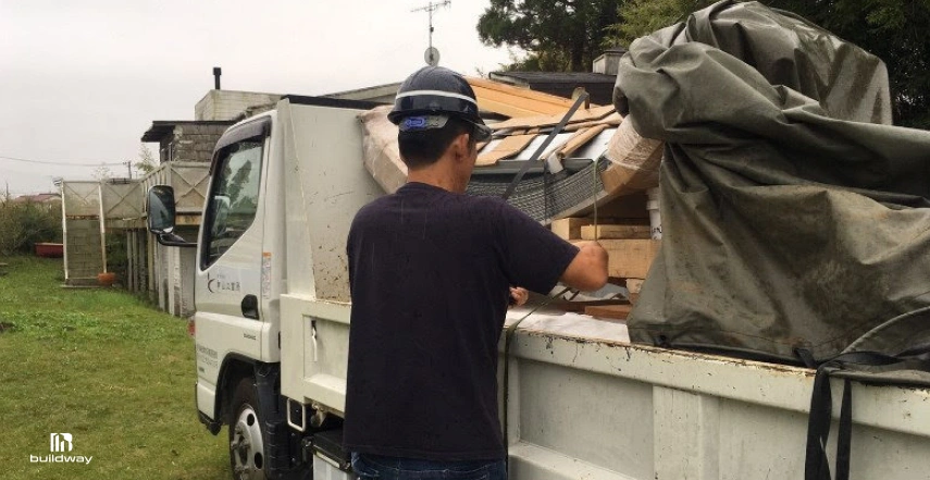 Construction worker in a hard hat inspecting a delivery truck loaded with building materials, including wooden pallets and covered supplies, at a residential or semi-rural site.