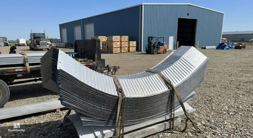 Stack of curved steel panels strapped for transport in front of a blue industrial building, with crates, forklifts, and trucks visible in the background.