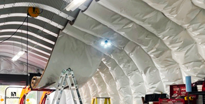 Interior of a Quonset-style steel building with white insulation panels being installed along the curved ceiling structure.