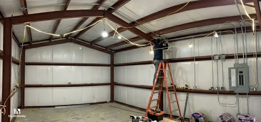 Person installing electrical wiring inside an insulated steel building with exposed red framing, conduit, and a visible breaker panel.