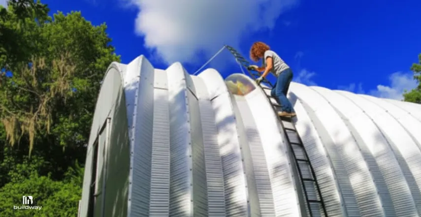 A woman on a ladder cleaning the curved metal roof of a building under a bright blue sky, surrounded by lush green trees.