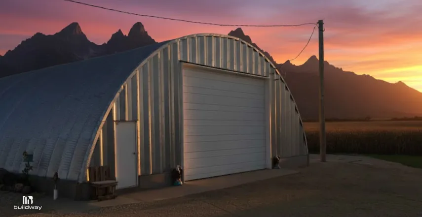 Quonset hut–style steel building with a curved metal roof and large roll-up door, set on a paved pad with mountains in the background at sunset.