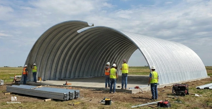 Construction crew assembling a curved steel Quonset-style building on a concrete slab in an open field.